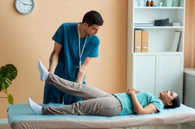 A physiotherapist guiding an elderly patient through a gentle knee-extension exercise at home, seated on a firm chair